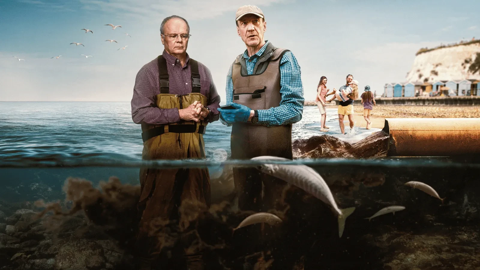Two people wearing protective clothing stand in shallow coastal water near an outflow pipe, with fish visible underwater and a beach scene behind them.