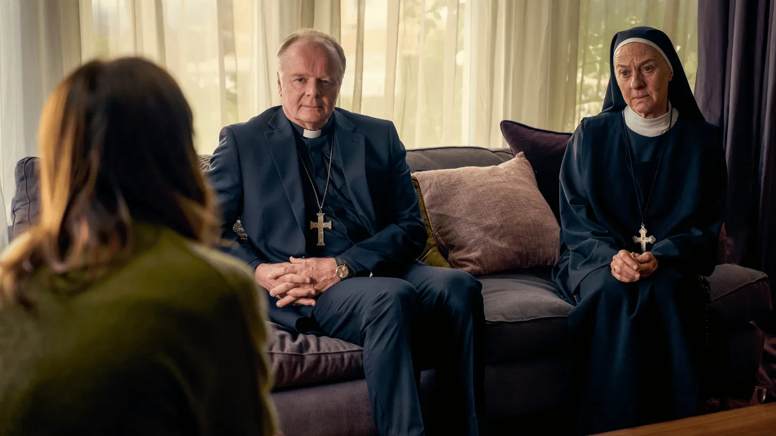 A priest and a person in a nun’s habit sit on a sofa in a softly lit room, facing someone off-camera, with light filtering through curtains behind them. 
