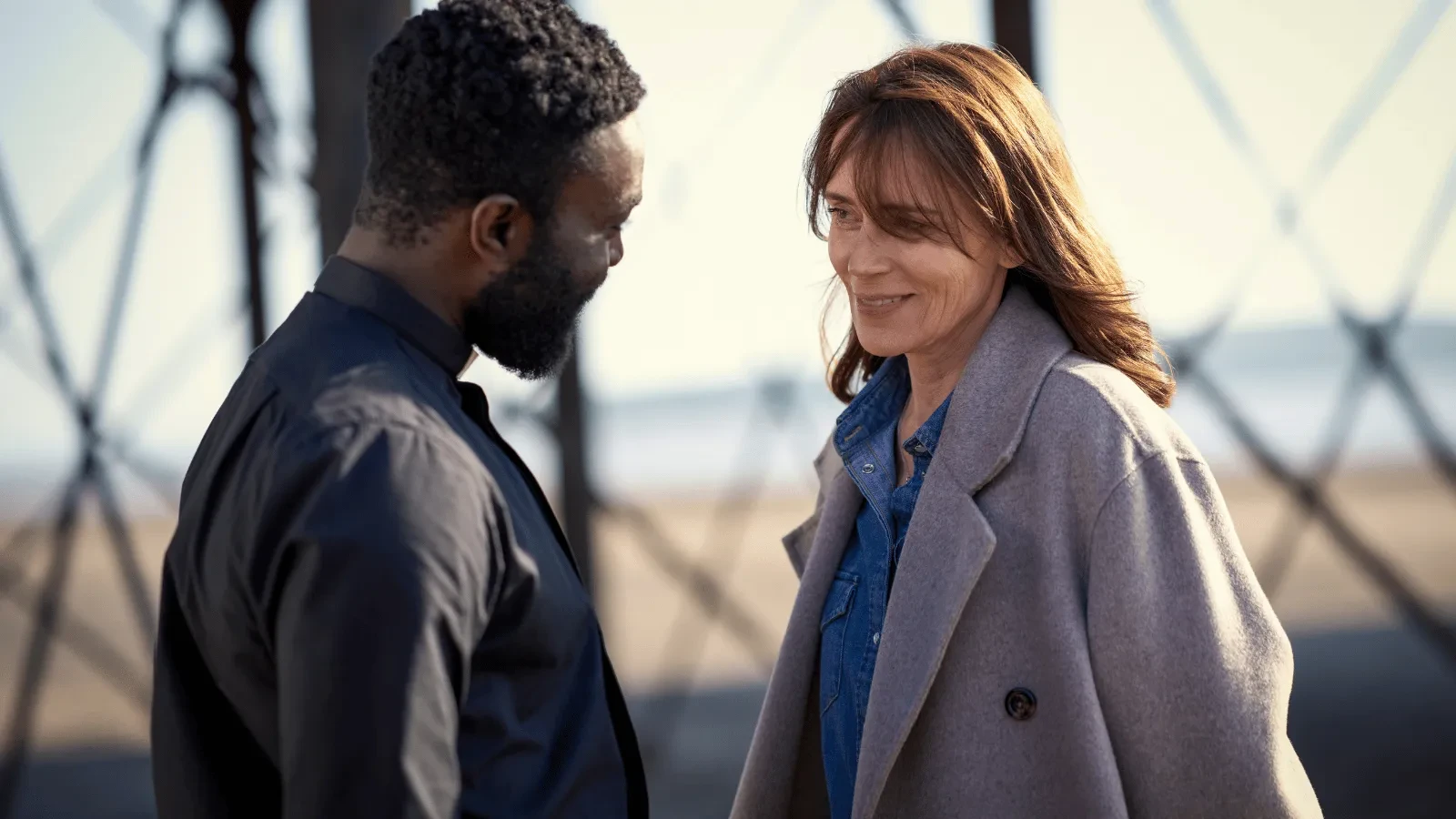 Two people stand close together on a seaside pier, facing each other in conversation, with blurred railings and water in the background. 