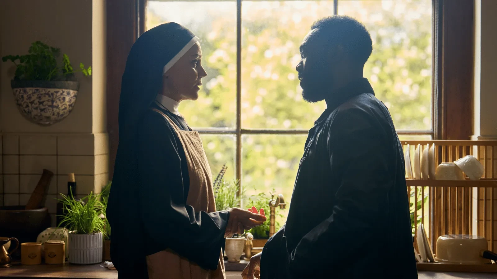 A person wearing a nun’s habit and apron stands face-to-face with a person in dark clerical clothing in a softly lit kitchen, with a window, plants, and dishes in the background.