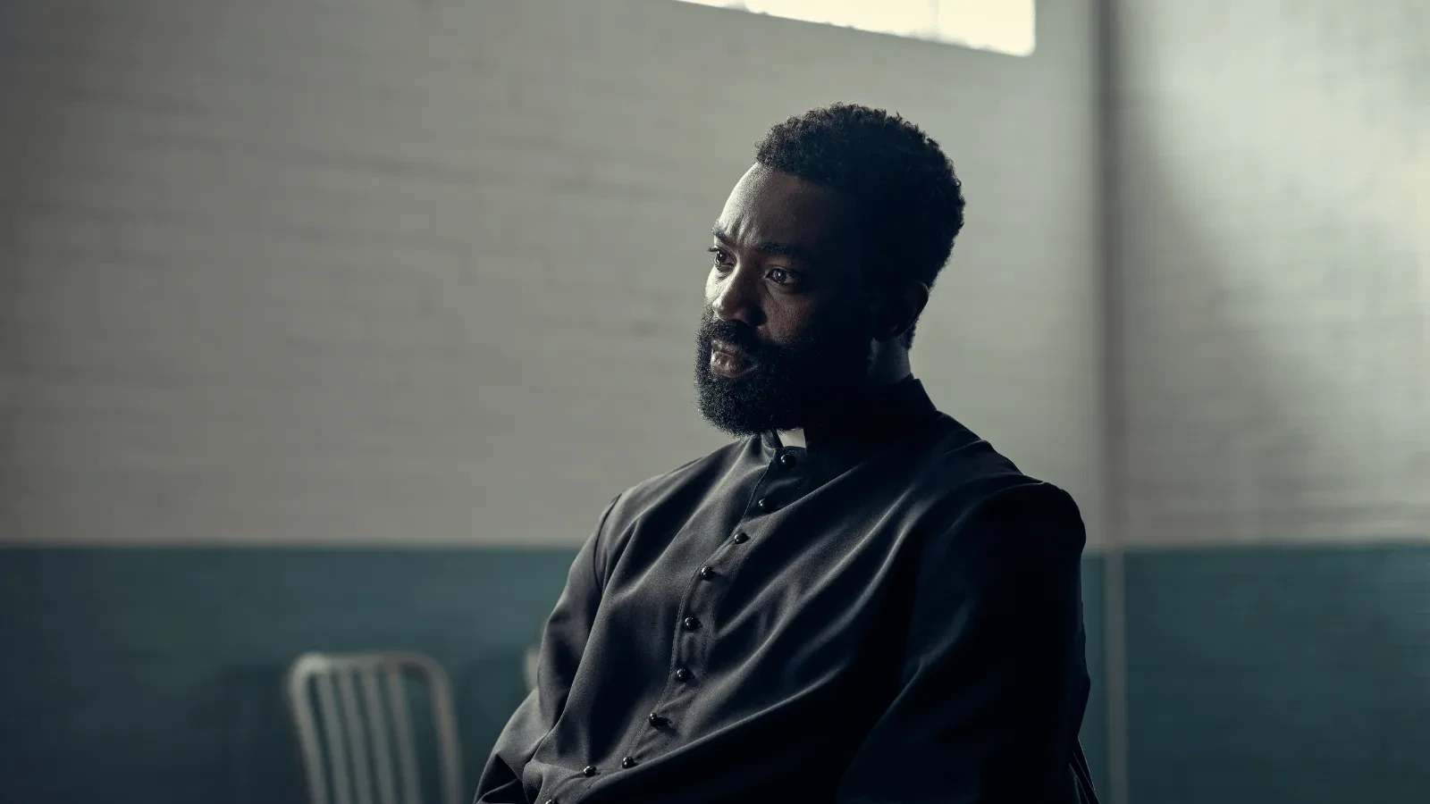  A person in priest’s clerical clothing sits indoors in a softly lit room, facing slightly to one side, with a plain wall and window behind. 