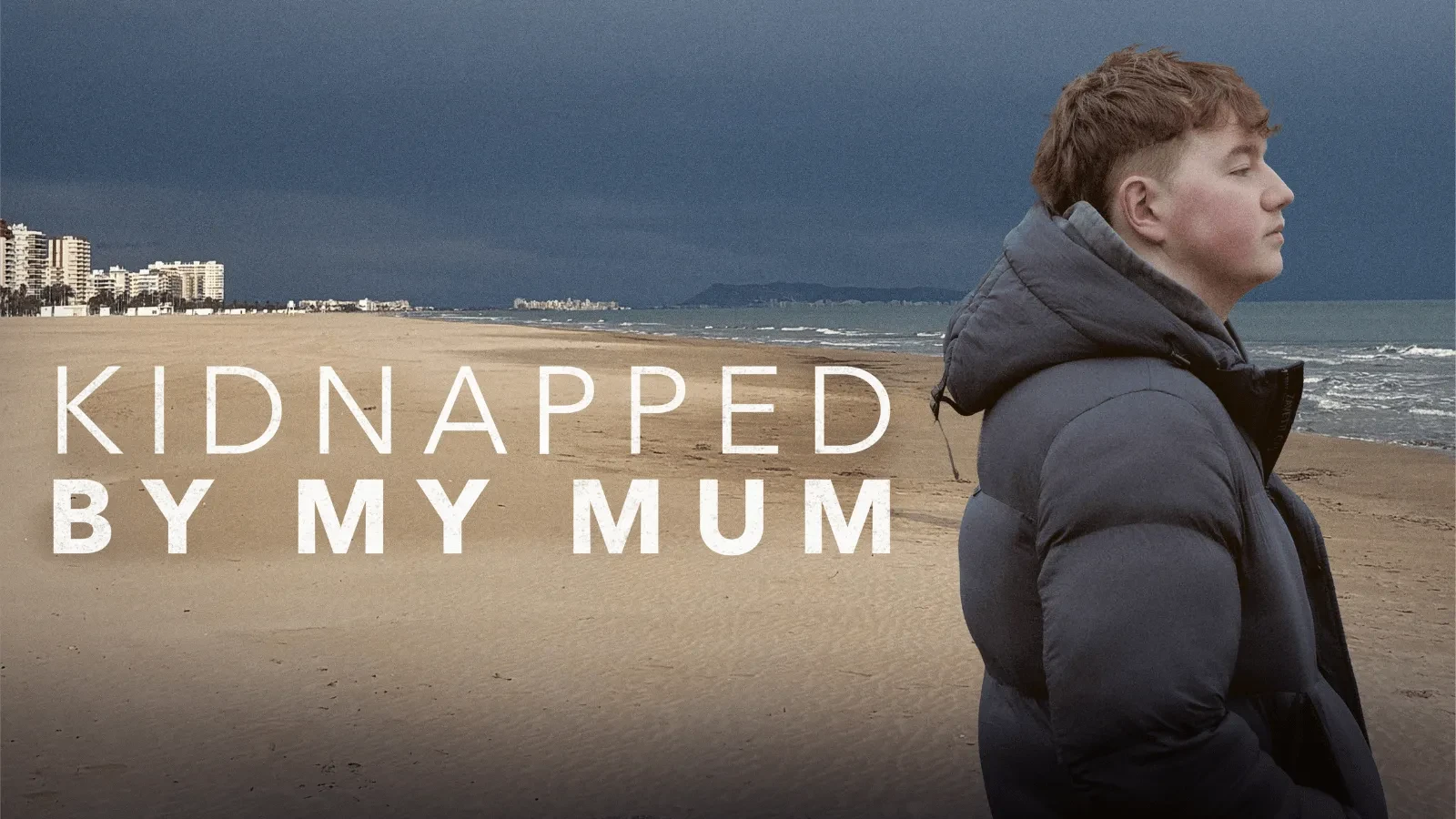 A Young Man wearing a puffer jacket stood on a beach with writing that reads kidnapped by my mum