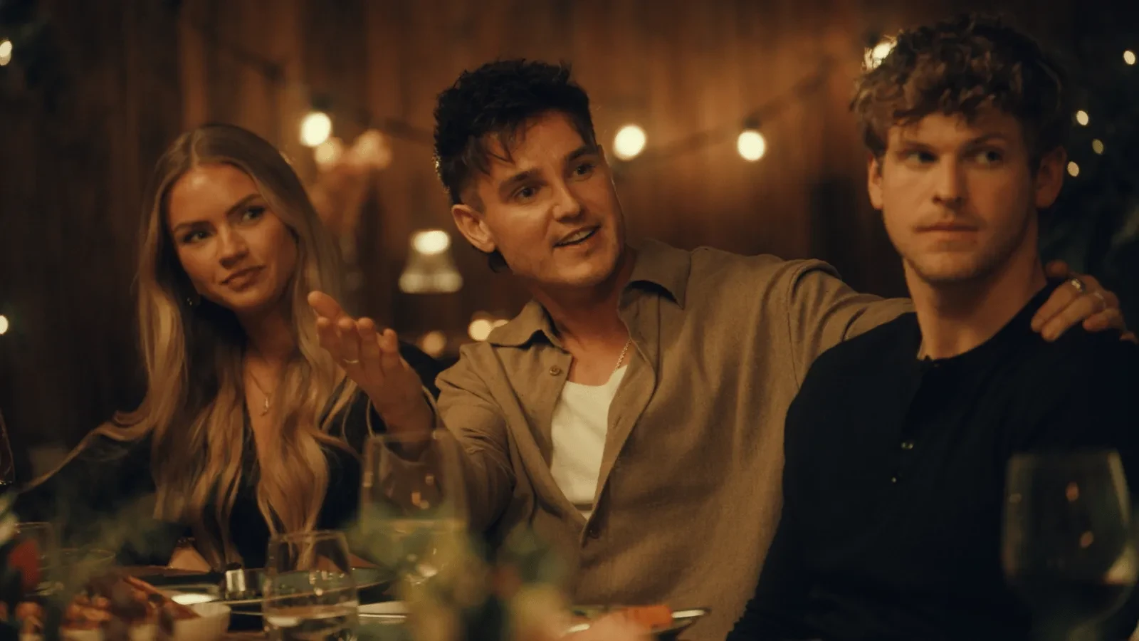 Three people seated at a dining table in a softly lit restaurant setting, talking while food and wine glasses sit in front of them.