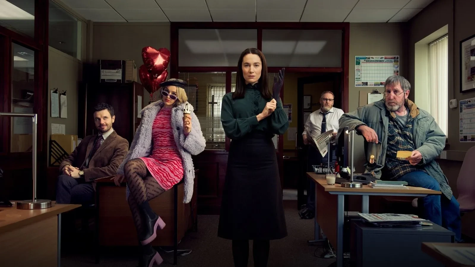Drama series promotional image set in a newsroom office, showing multiple people positioned around desks with newspapers, paperwork, and office lighting.