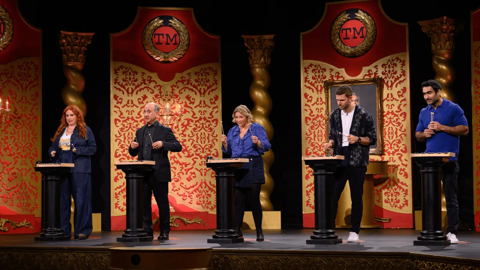 Five people standing at individual podiums on a stage, each holding small mallets, with ornate red-and-gold set pieces behind them.