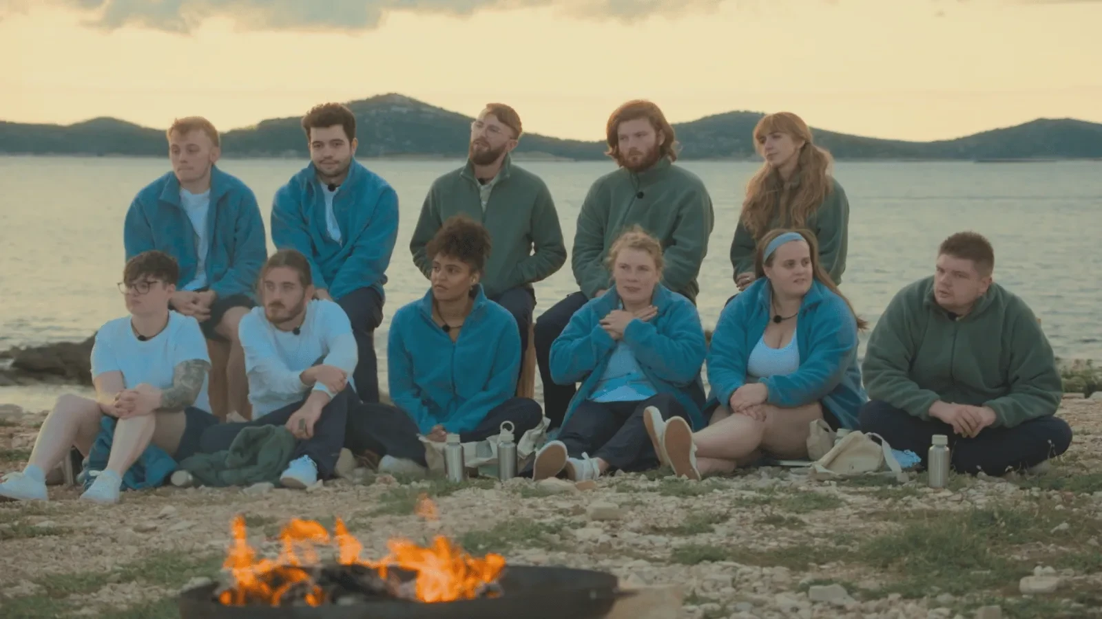 A group of people sitting together outdoors near a small fire, wearing matching jackets, with water and hills behind them.