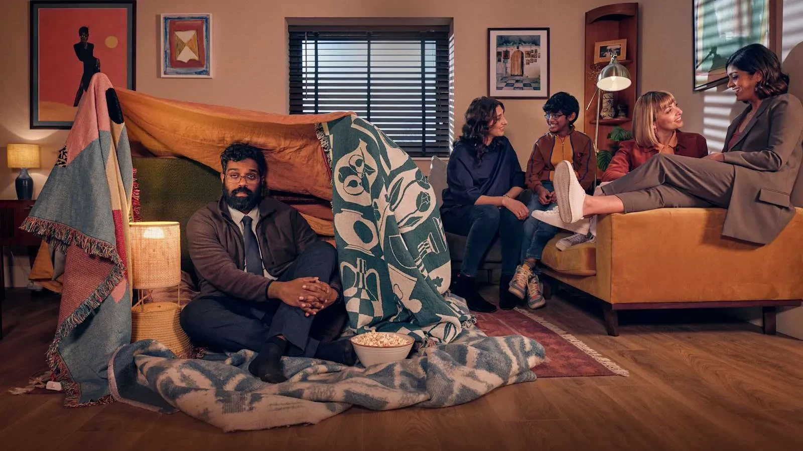 Romesh Ranganathan sat in a living room under a fort of blankets with a family sat in the background smiling. Key art for the show Avoidance.
