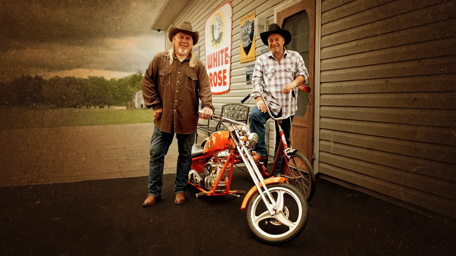 Two vintage-loving pickers stand outside a rustic shop with retro signs, showing off a bright custom motorbike and a tricycle find, surrounded by countryside charm.&nbsp;