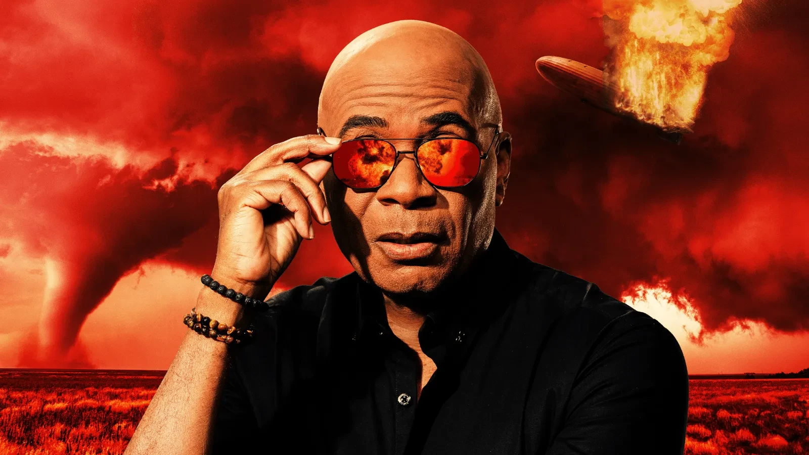 A person wearing reflective red sunglasses looks ahead against a dramatic red sky while a blazing airship explodes in the background over an open landscape. 