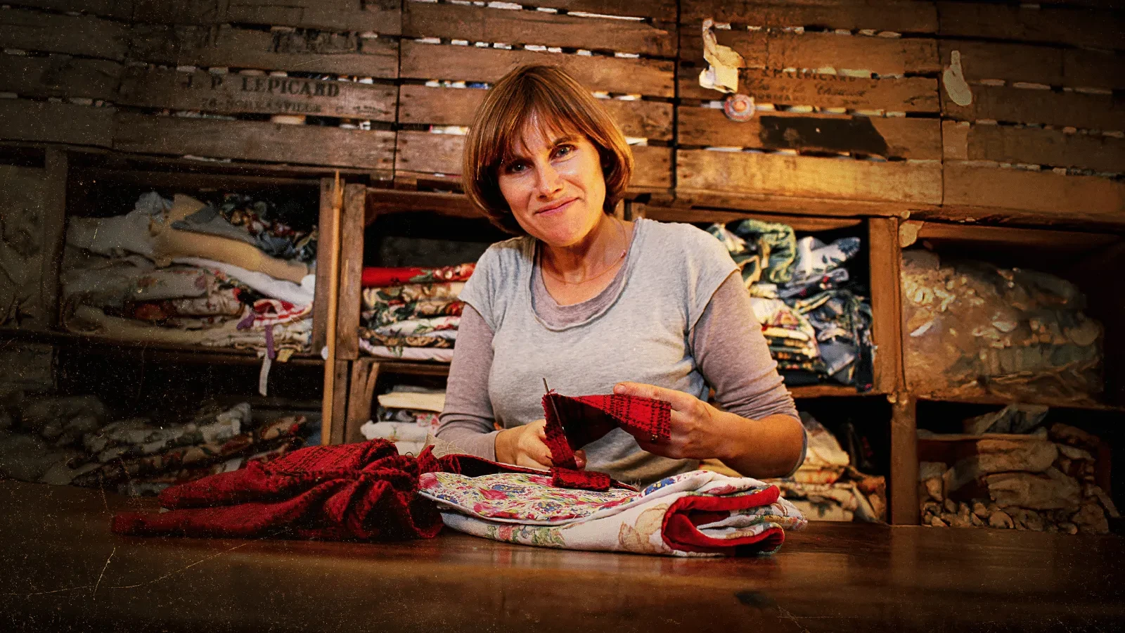 A furniture upcycler works at a wooden table, stitching colourful fabric in a packed workshop filled with neatly stacked textiles.