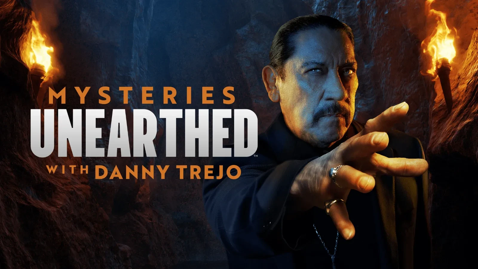 A person reaches forward inside a dimly lit cave setting, flanked by flaming torches, alongside large on-screen text reading “Mysteries Unearthed with Danny Trejo.” 