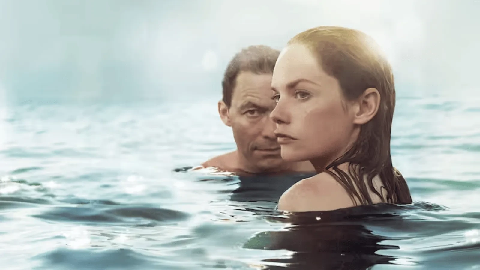 Two people are partially submerged in open water, with only their heads and shouldCers visible above the surface. The person in the foreground with slicked-back hair looks off to the right, while another person behind them looks forward.&nbsp; 
