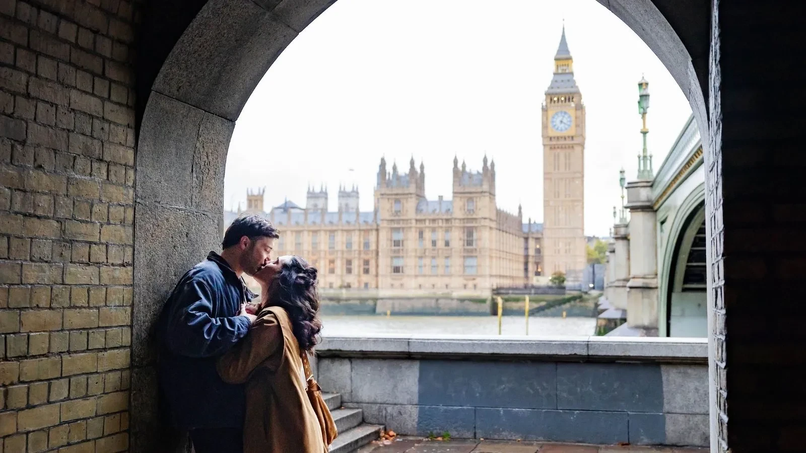 A young couple kissing under a tunnel with the Houses of Parliament in the background behind them 