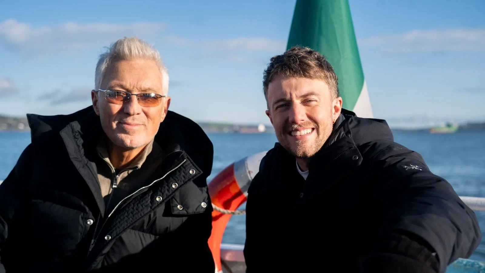 Martin Kemp and Roman Kemp stood on a boat in warm clothing with an Irish flag behind them and the sea in the background