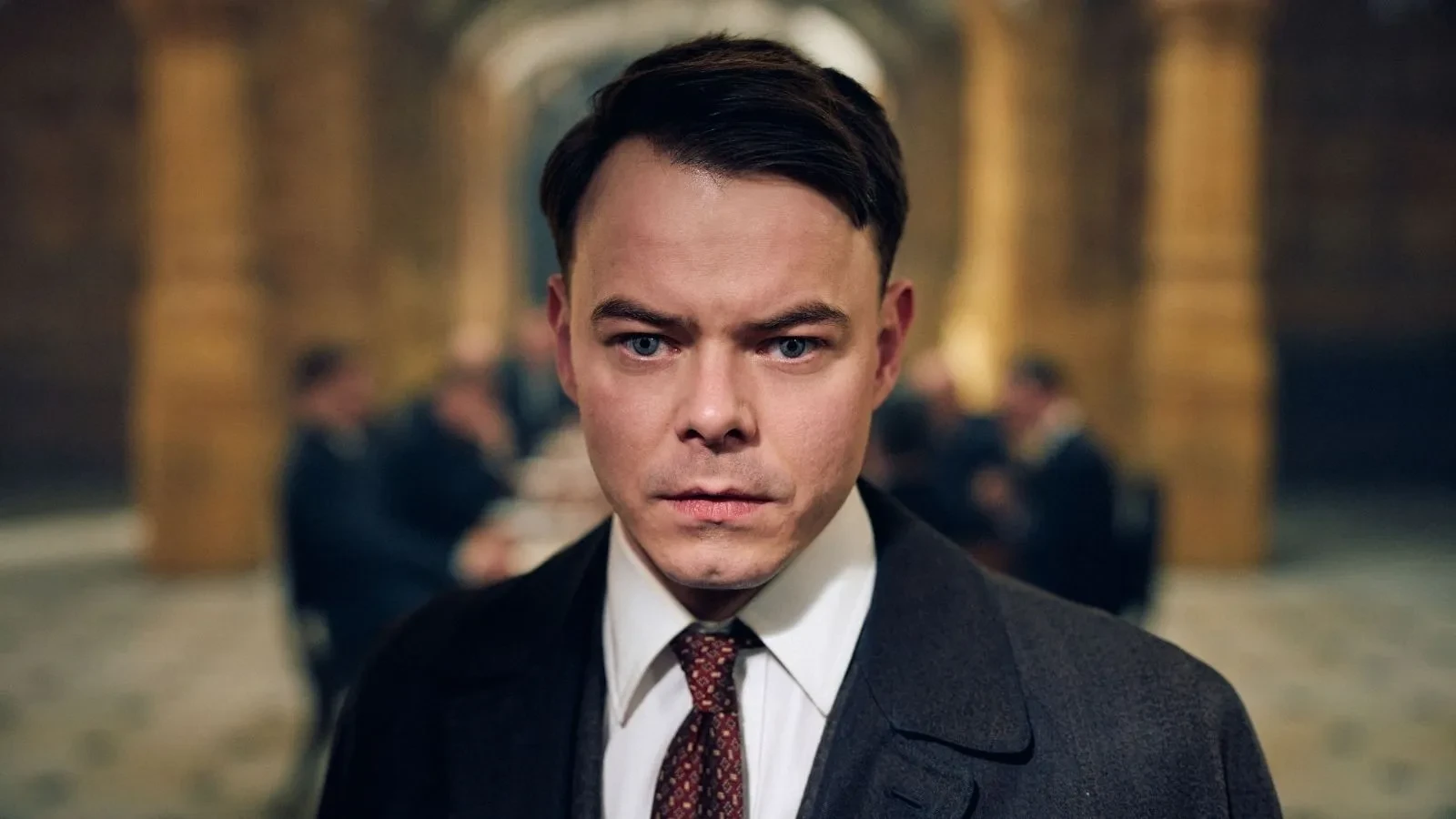 A man with dark hair in a suit with a blurred background of people sat behind him in a grand building