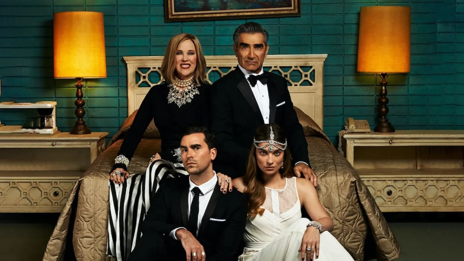Four people dressed in formal attire are posed in a hotel-style bedroom. Two figures sit on the bed while two stand behind them, framed by matching bedside lamps and teal-coloured walls.