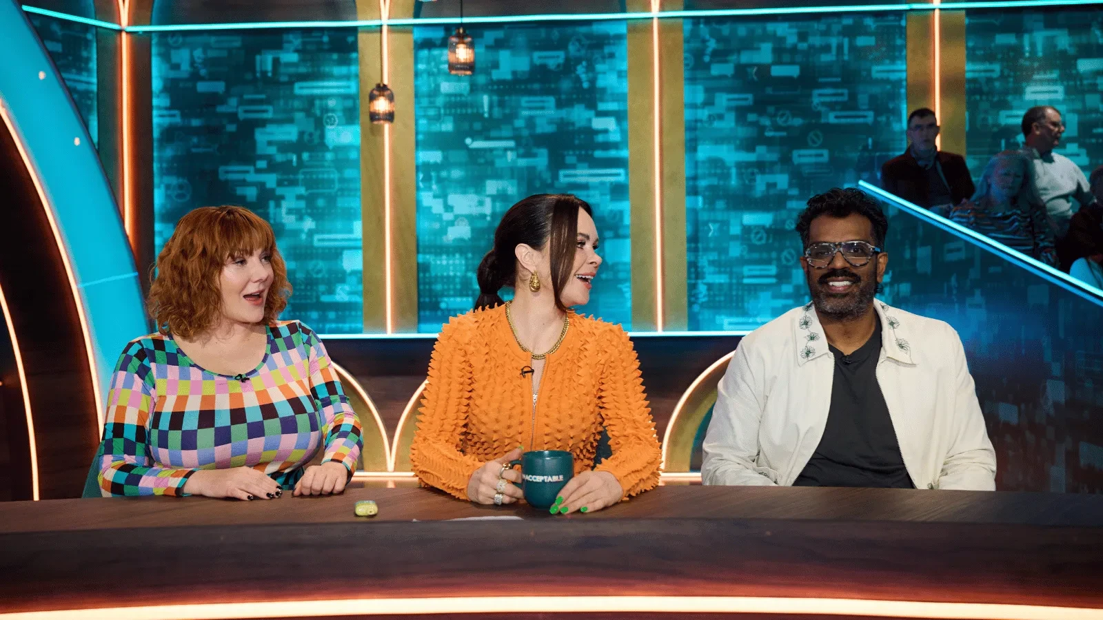  Three panelists seated behind a desk on a television studio set with blue digital-themed screens and neon lighting. One person wears a green headscarf, another a dark blazer, and a third a sparkling blue jacket.