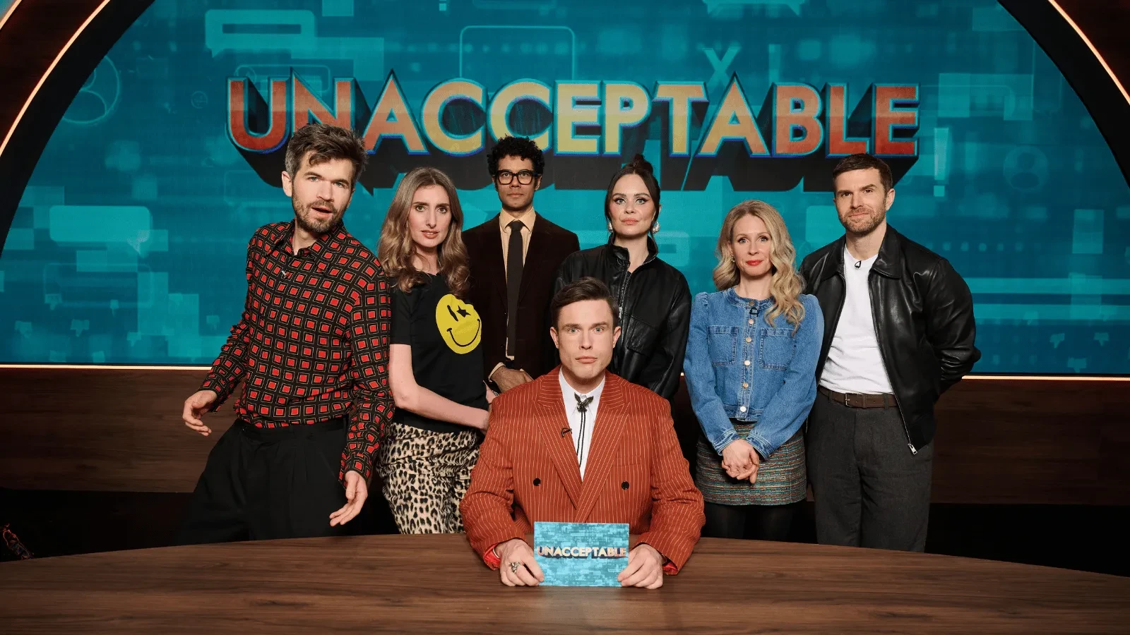Six people pose on a television studio set in front of a large “UNACCEPTABLE” sign. One person sits at a desk holding a programme card while five others stand behind, framed by blue graphics and warm studio lighting. 