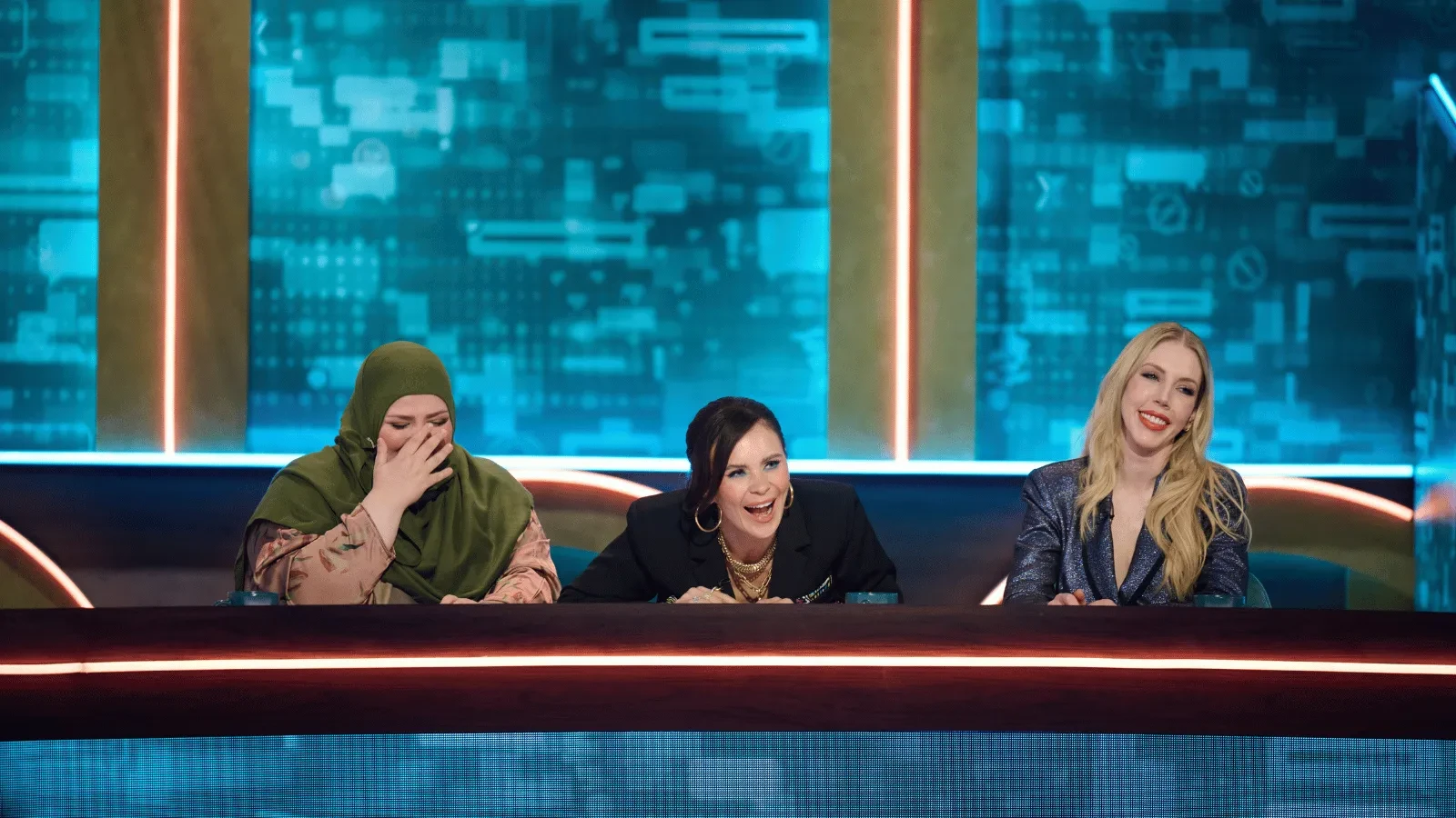 Three panelists seated behind a curved desk on a brightly lit studio set with blue digital-style graphics. One person in a colourful checkered top, one in a textured orange jacket holding a mug, and one in a light-coloured jacket are engaged in conversation. 