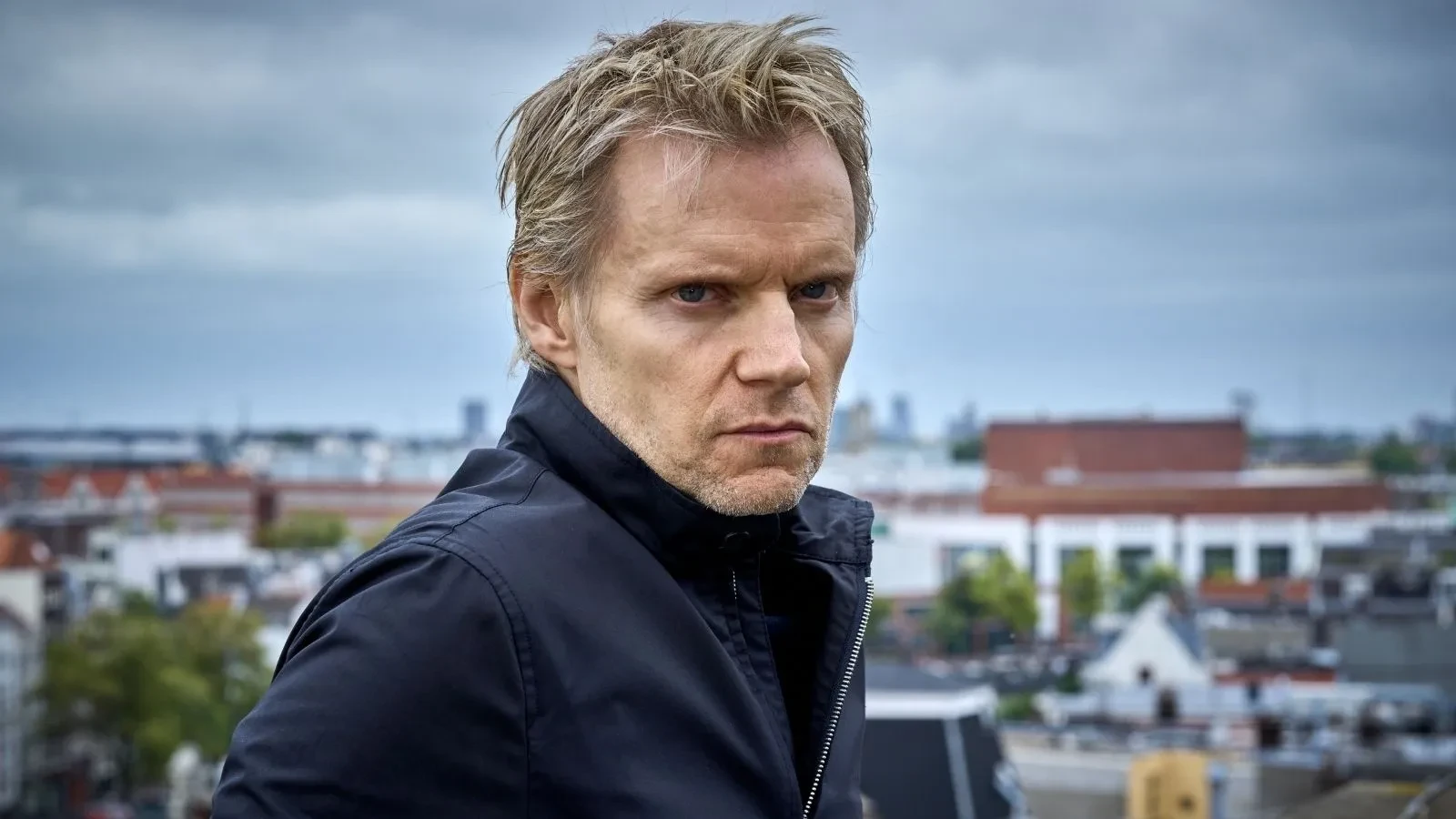 A man in his forties stood on a roof top wearing a black jacket looking straight into the camera with his buildings behind him.