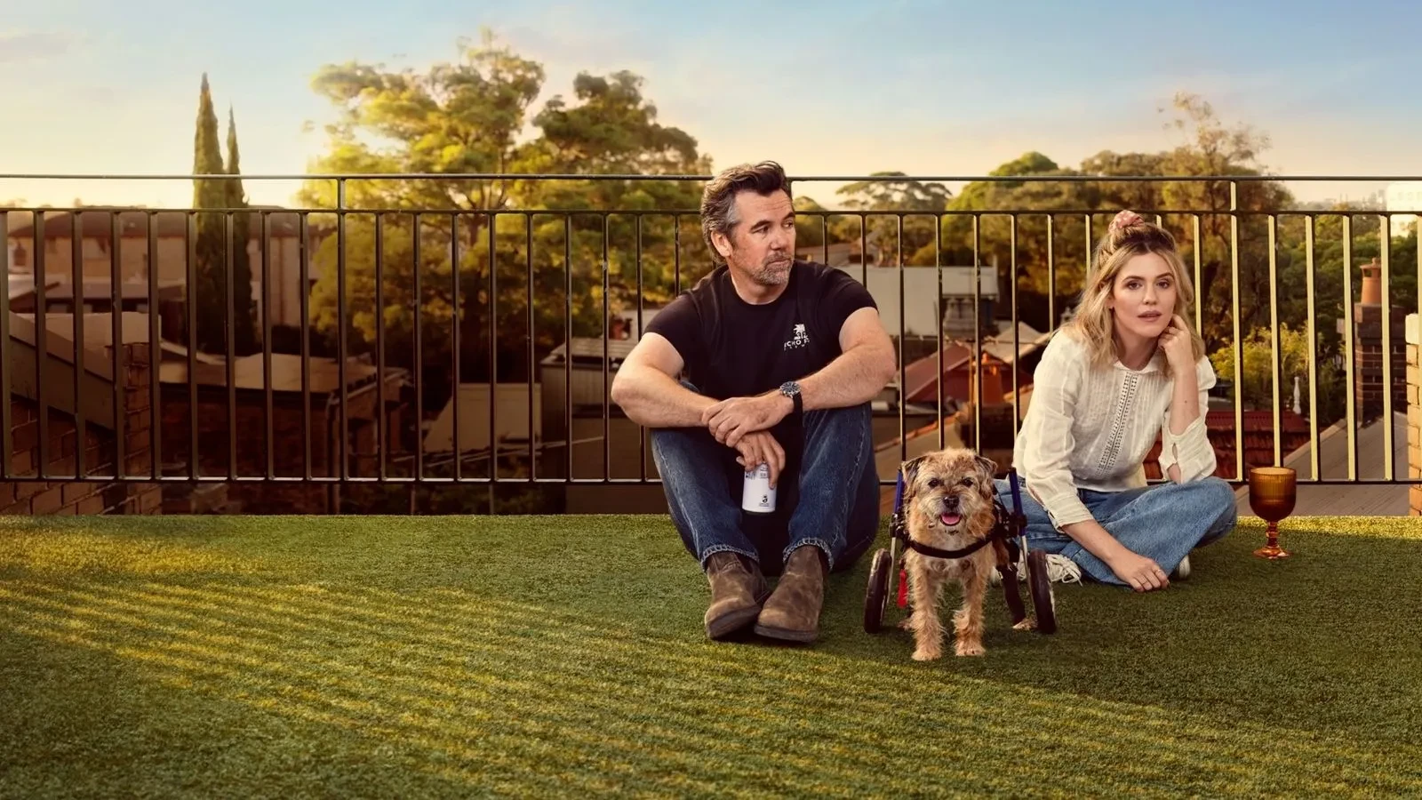 A man and a woman sat on a balcony with astro turf with a dog in a wheelchair sat between them 