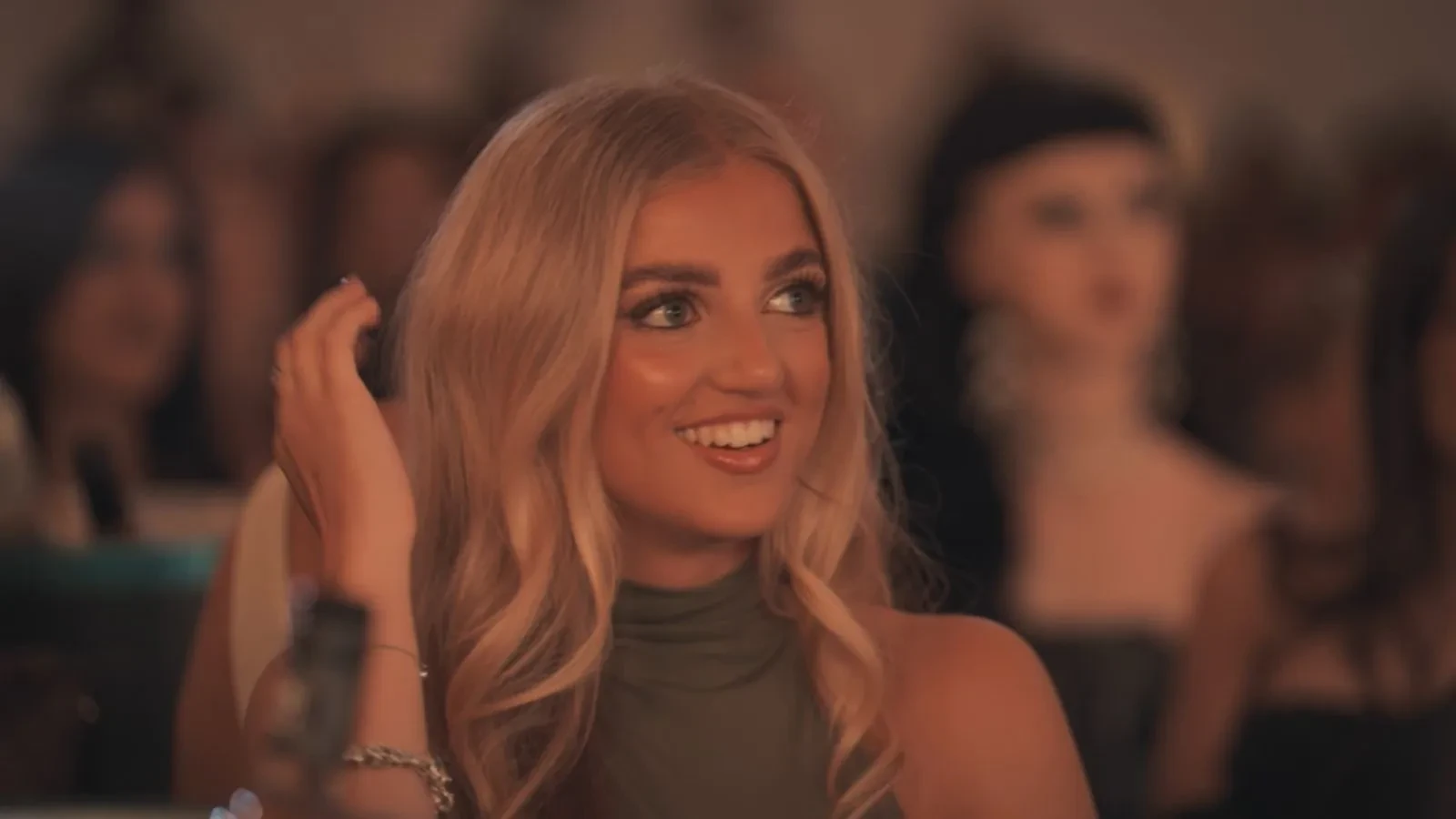 A close-up promotional image showing a person seated at an event or party under warm lighting. The person is surrounded by a blurred crowd in the background, creating a glamorous reality TV atmosphere with guests gathered around tables.