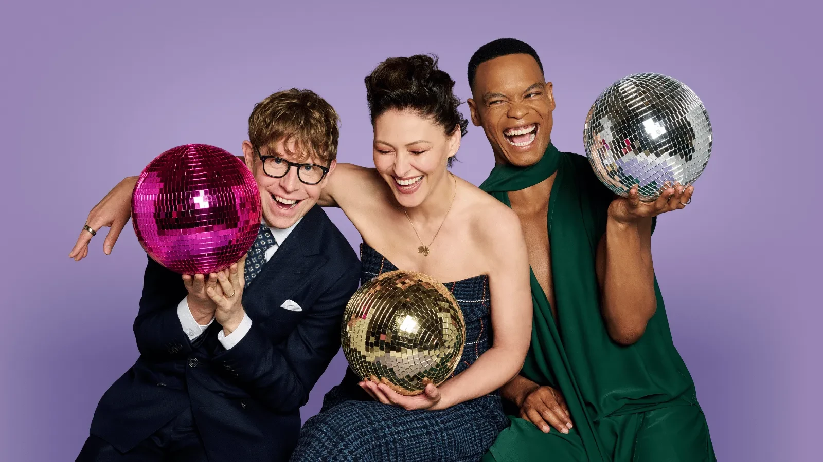 Three presenters pose against a purple backdrop while holding colourful disco balls. The bright studio image captures the sparkle and glamour associated with the Strictly Come Dancing ballroom.