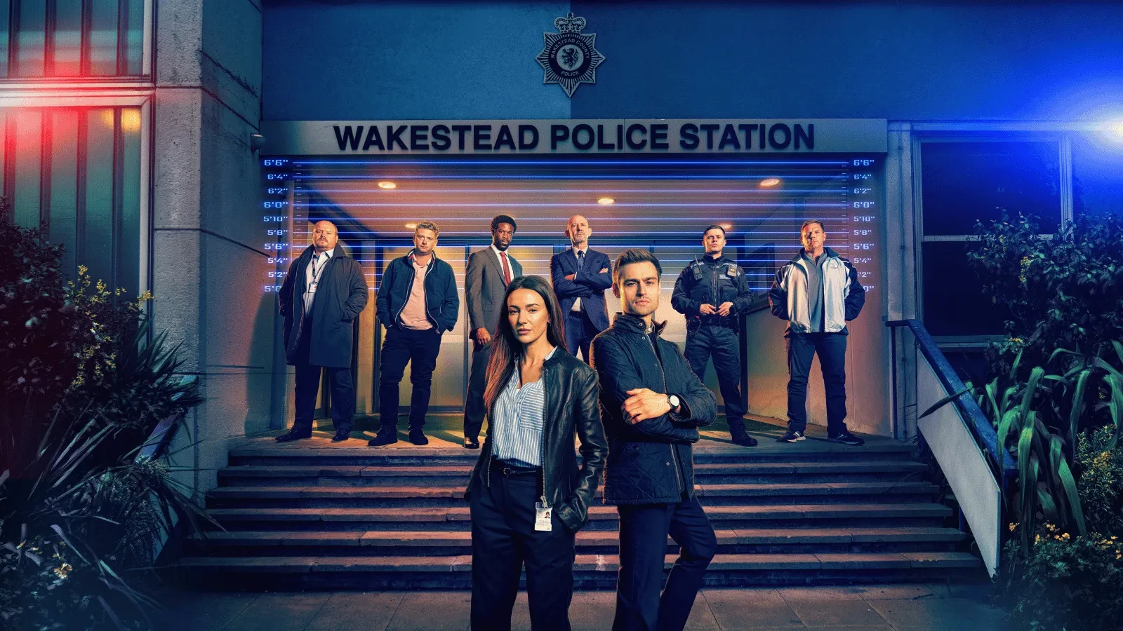 Promotional cast image for a crime drama showing a group of detectives and police officers standing outside Wakestead Police Station at night. Two lead characters stand in the foreground, while the wider team gathers on the station steps behind them, illuminated by blue and red police lights.