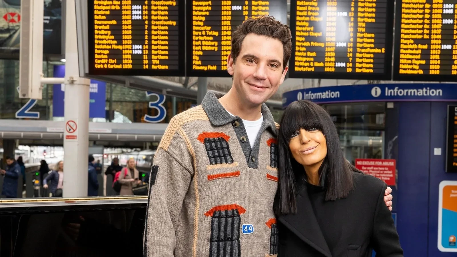 Two presenters stand together inside a busy train station beneath large departure boards displaying travel information. The promotional image for The Piano features the pair on a station platform, highlighting the show's journey across the UK to discover talented amateur pianists.