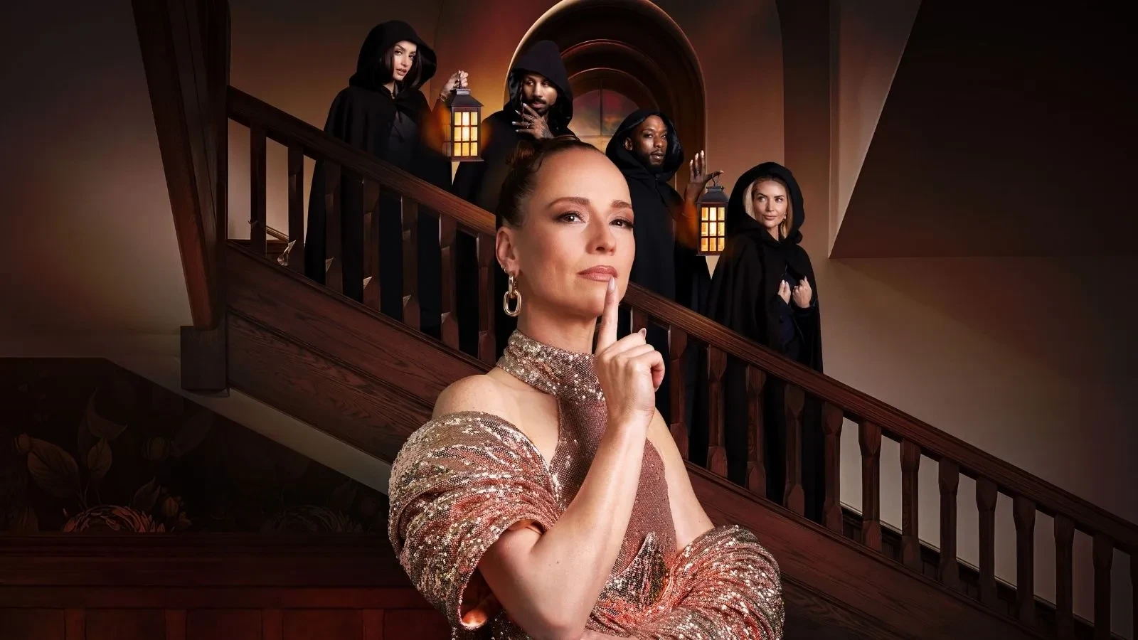 A promotional image for The Traitors Canada featuring a person in a shimmering gold outfit standing in front of a grand wooden staircase, holding a finger to their lips. Several hooded contestants stand on the staircase behind them carrying lanterns, creating a mysterious atmosphere inside a historic manor setting.  