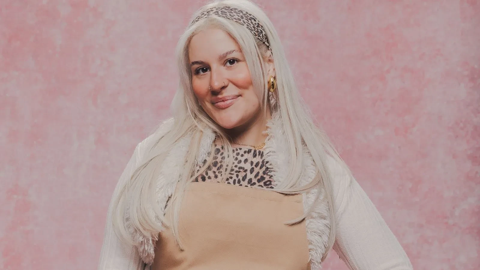 Great British Bake Off baker Clara posing in a baking apron against a pink studio background.
