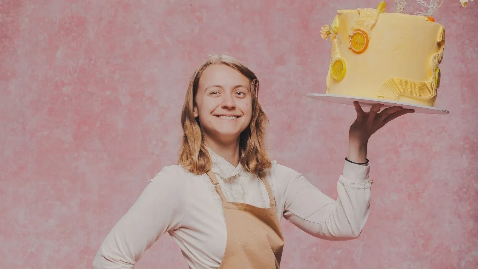 Great British Bake Off baker Danni holding a decorated yellow citrus cake against a pink studio backdrop.