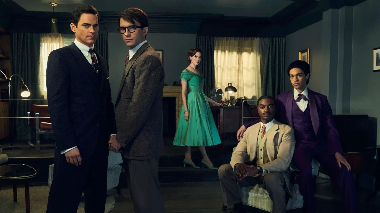 Two young men in suits holding hands with another couple behind them and a women in a 1940s style dress  in  an old living room.