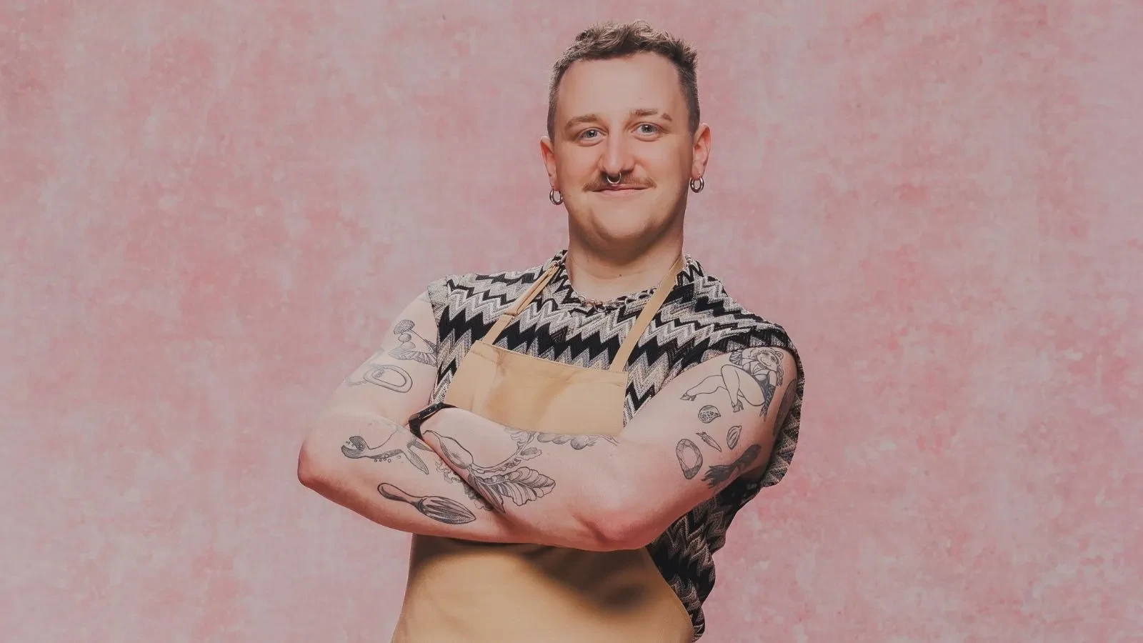 Great British Bake Off baker Gabe wearing a baking apron and patterned top in an official GBBO contestant portrait.