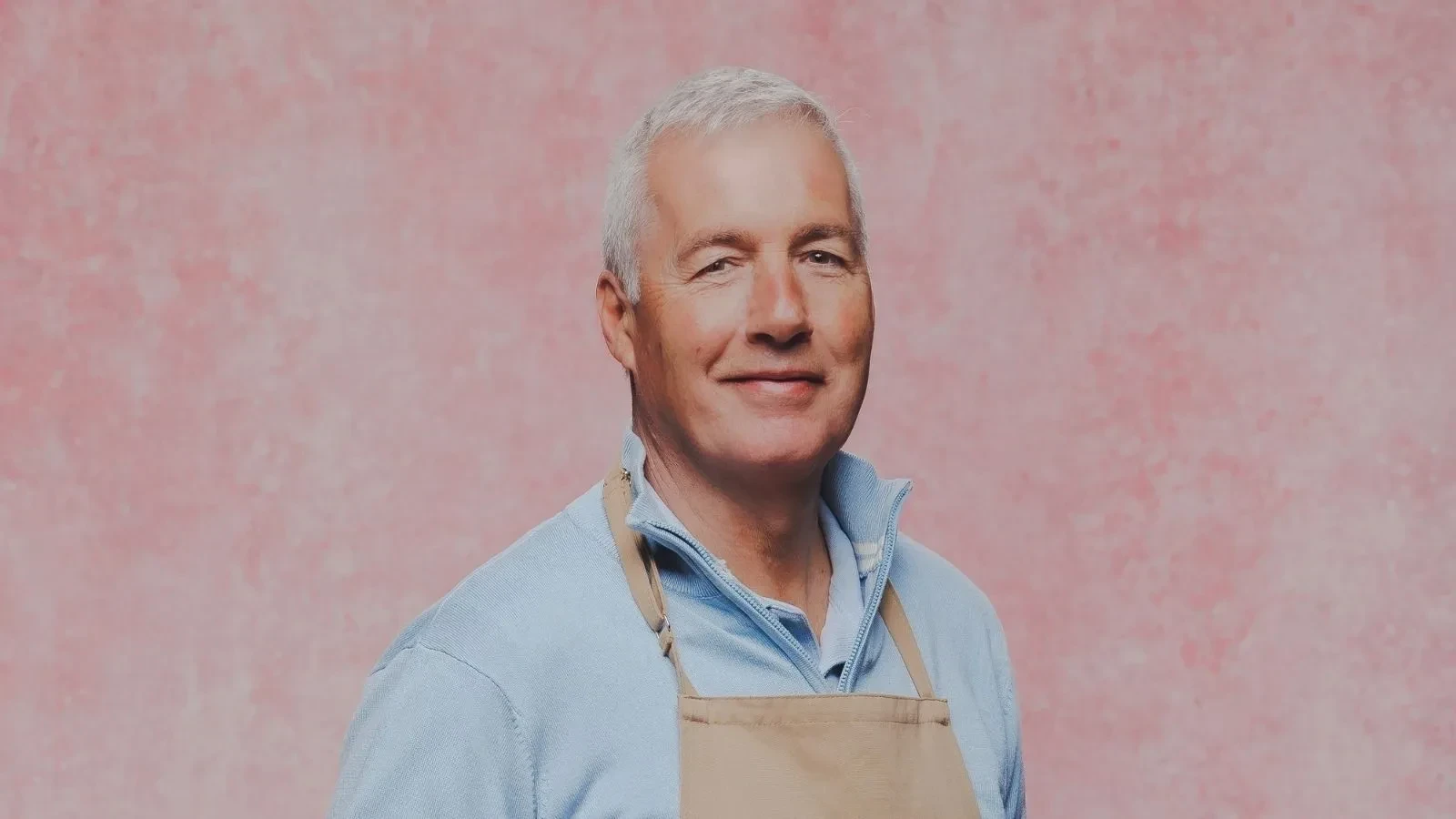 Great British Bake Off baker Gary wearing a baking apron in an official GBBO contestant portrait against a pink studio backdrop.