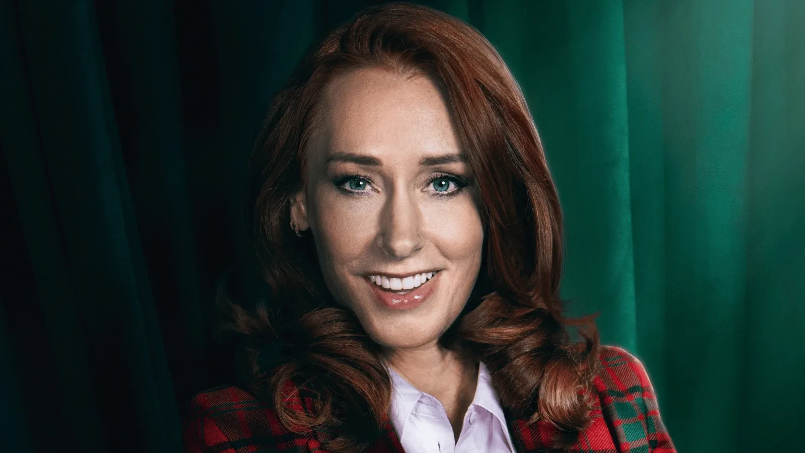 Close-up studio portrait of a person with long auburn hair, wearing a red tartan blazer over a light-coloured shirt. The subject faces the camera against dark green curtains, with soft lighting highlighting facial features and hair.