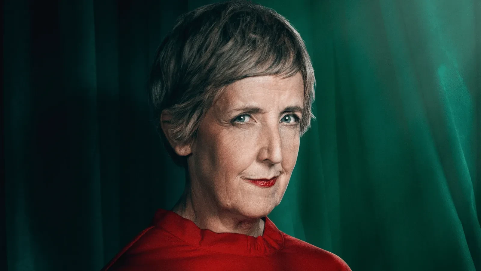 Close-up studio portrait of a person with short hair, wearing a bright red top. The subject is positioned in front of dark green curtains, with focused lighting highlighting facial features and contrasting with the vivid clothing.