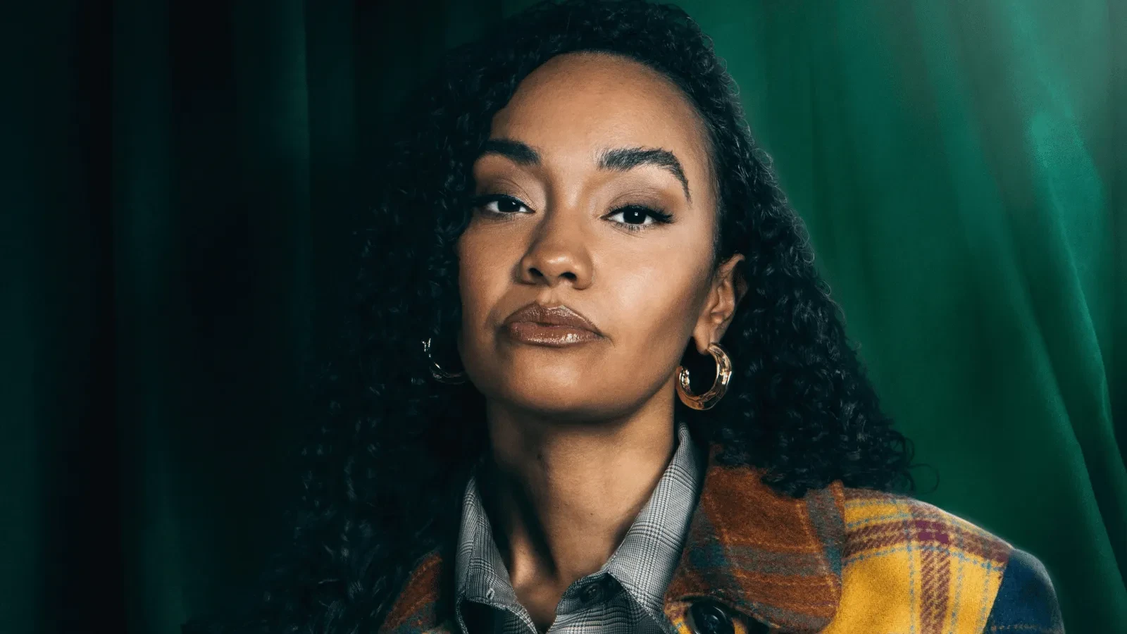 Close-up studio portrait of a person with curly hair, wearing hoop earrings and a patterned coat over a collared shirt. Dark green curtains and directional lighting create a rich, textured background.