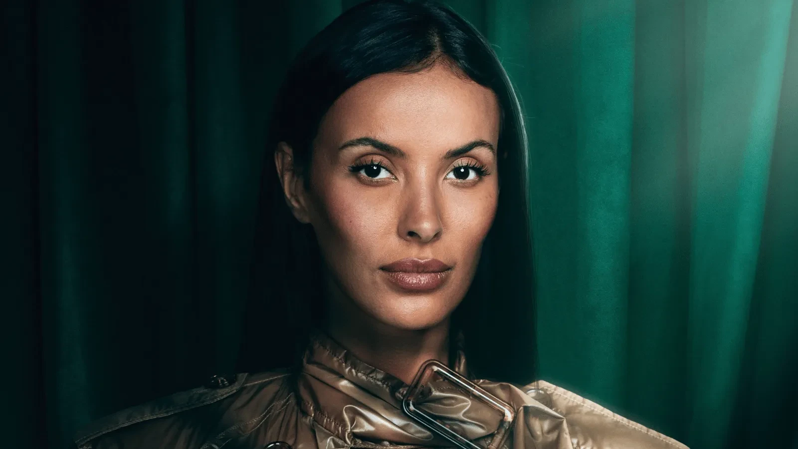Close-up studio portrait of a person with long dark hair, wearing a metallic high-neck jacket. The subject faces the camera against a backdrop of deep green curtains, lit with soft, dramatic lighting.