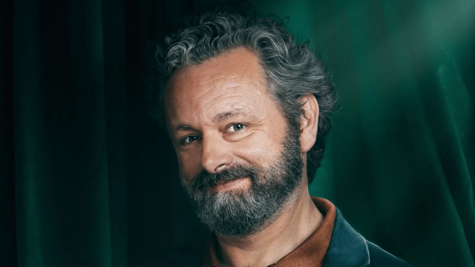 Close-up studio portrait of a person with wavy grey hair and a beard, wearing a collared top and jacket. The subject is positioned in front of deep green curtains, illuminated by directional lighting that creates contrast and depth.
