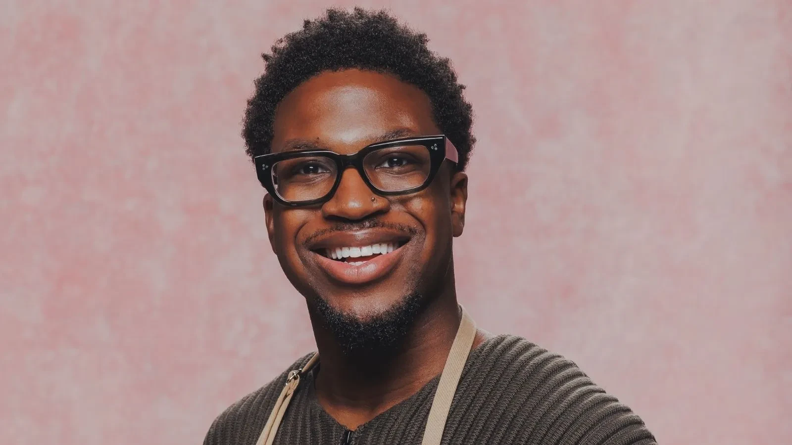 Great British Bake Off baker Moyin wearing glasses and a baking apron in an official GBBO contestant portrait.