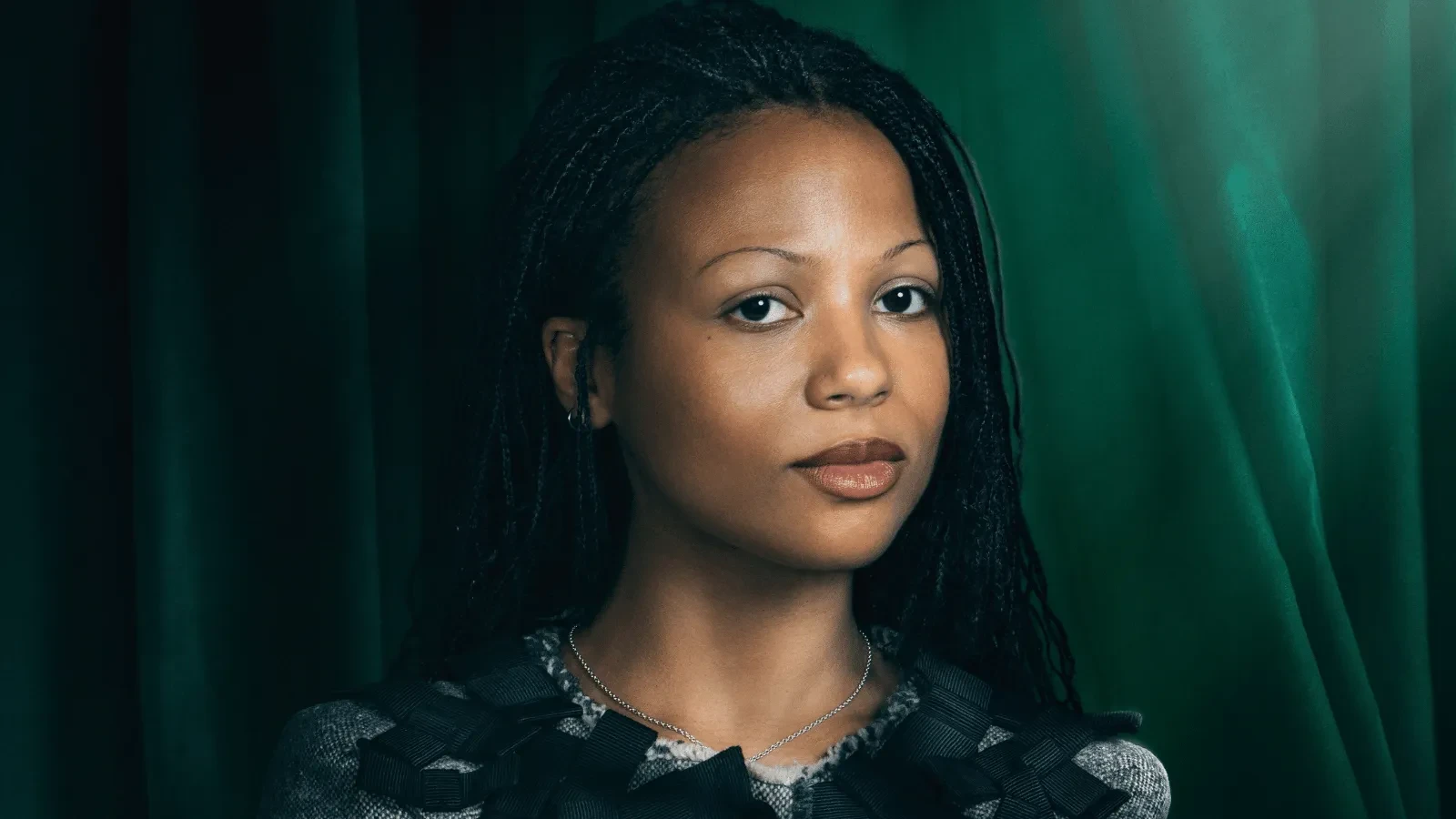 Close-up studio portrait of a person with long braided hair, wearing a textured top with ribbon detailing and a delicate necklace. The subject is photographed against dark green curtains with soft, dramatic lighting and a clear, uncluttered composition.