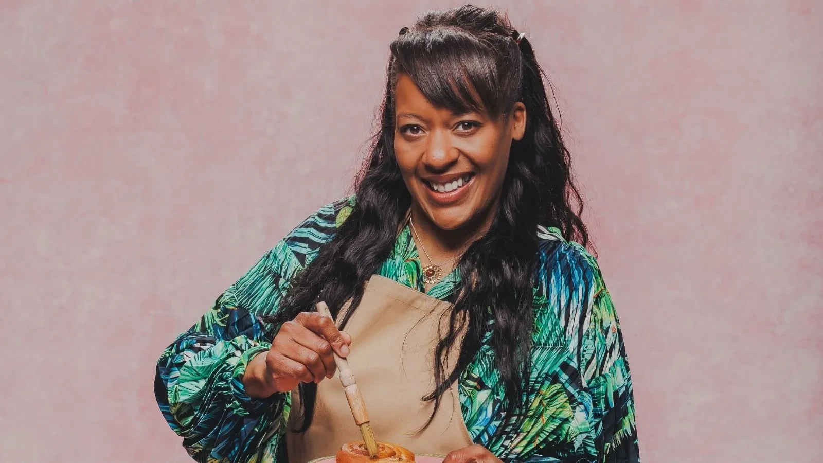 Great British Bake Off baker Nikki holding a pastry brush over a baked pastry in an official GBBO contestant portrait.