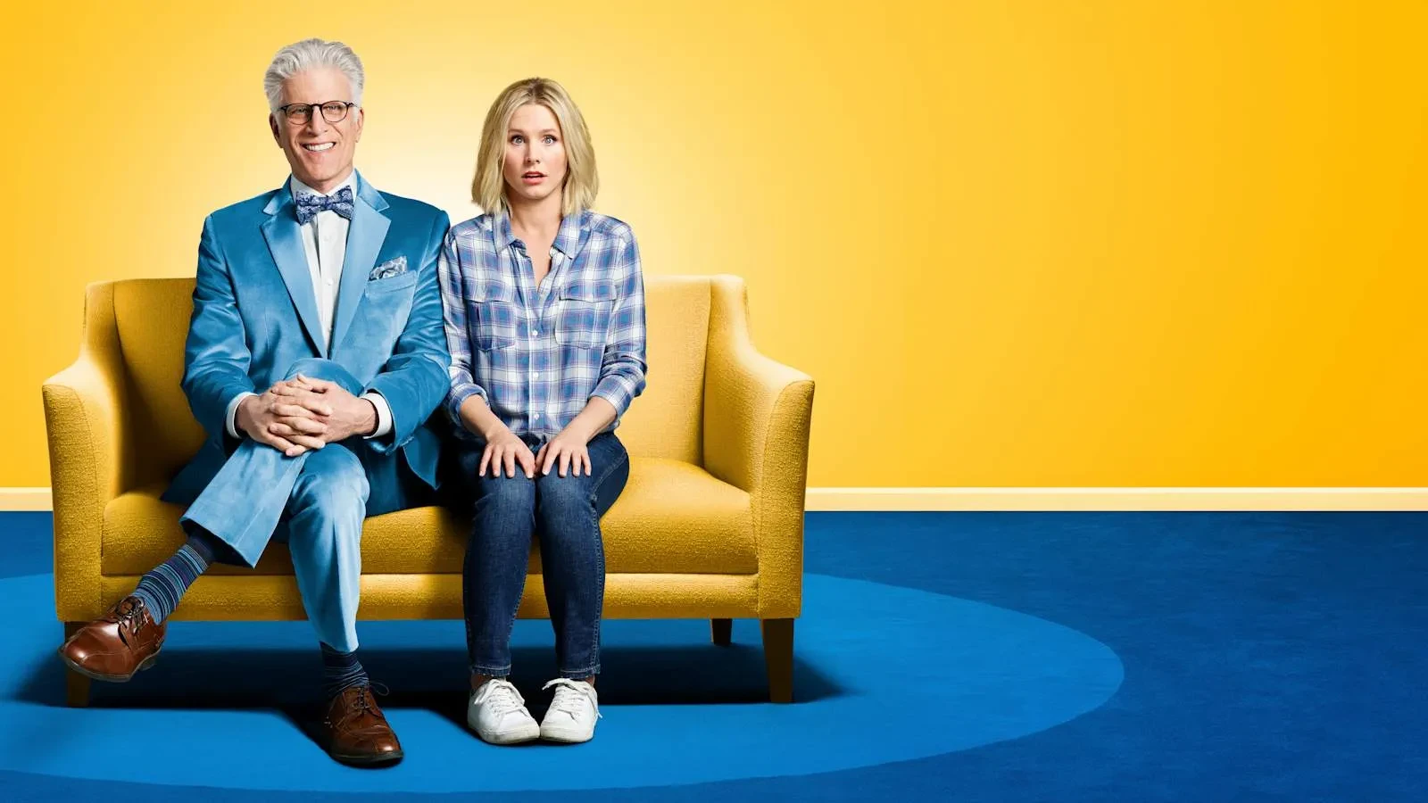 A man and women sat on a yellow sofa in a yellow room with a blue carpet. Key art for The Good Place.