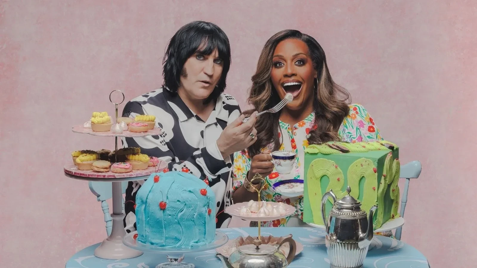 Great British Bake Off hosts gathered around a table with cakes, pastries and afternoon tea treats in an official GBBO promotional image.