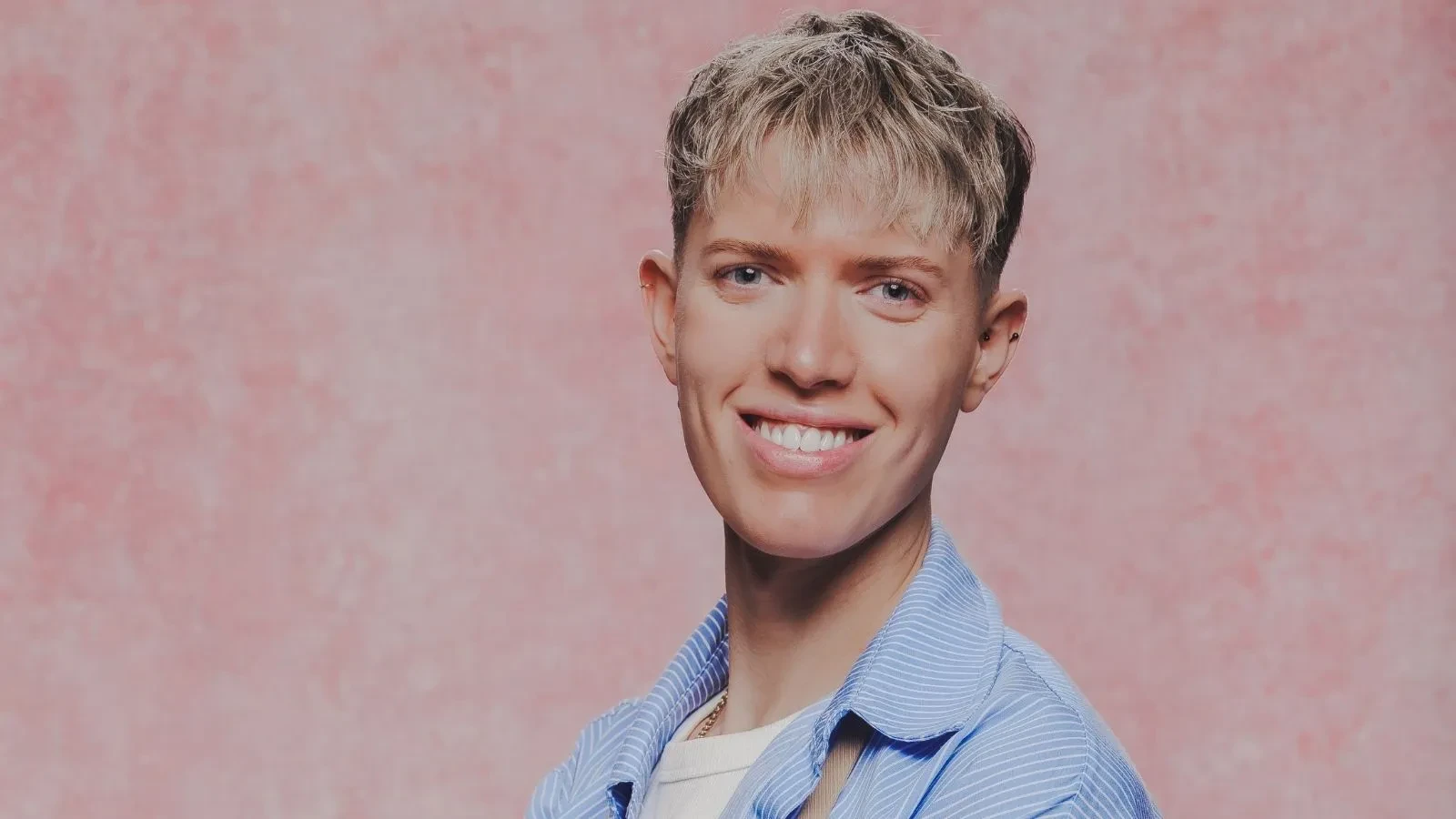 Great British Bake Off baker Tom in an official GBBO contestant portrait against a pink studio backdrop.