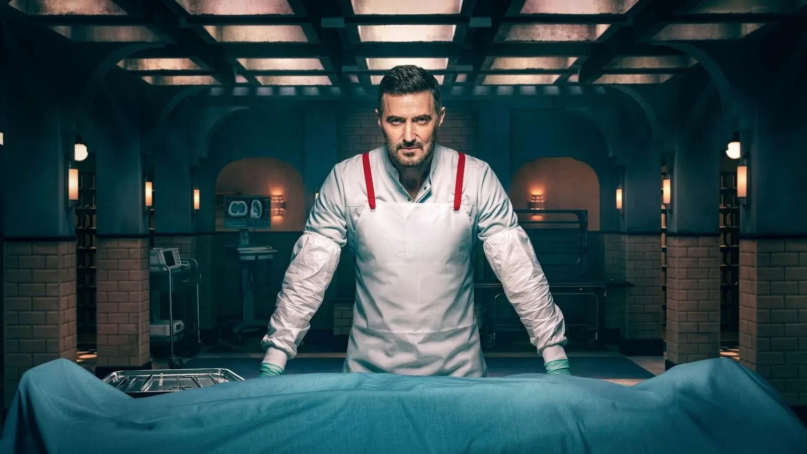 Richard Armitage stood in a morgue in front of a body covered in a blue blanket. Key art for ITVs new drama Winter.