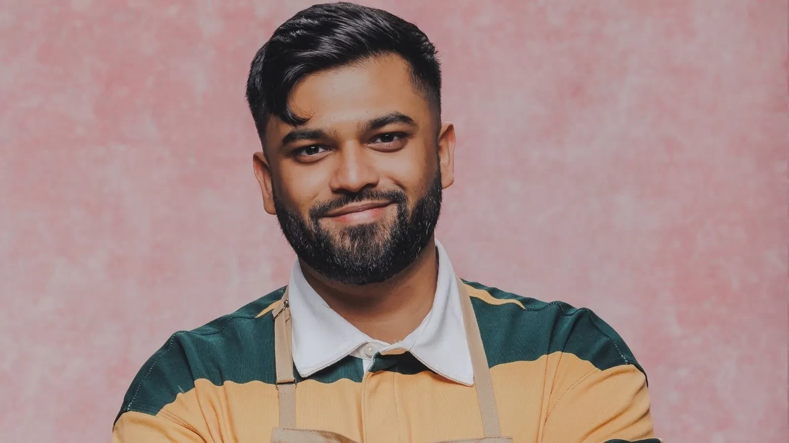 Great British Bake Off baker Yannis wearing a baking apron in an official GBBO contestant portrait against a pink studio backdrop.