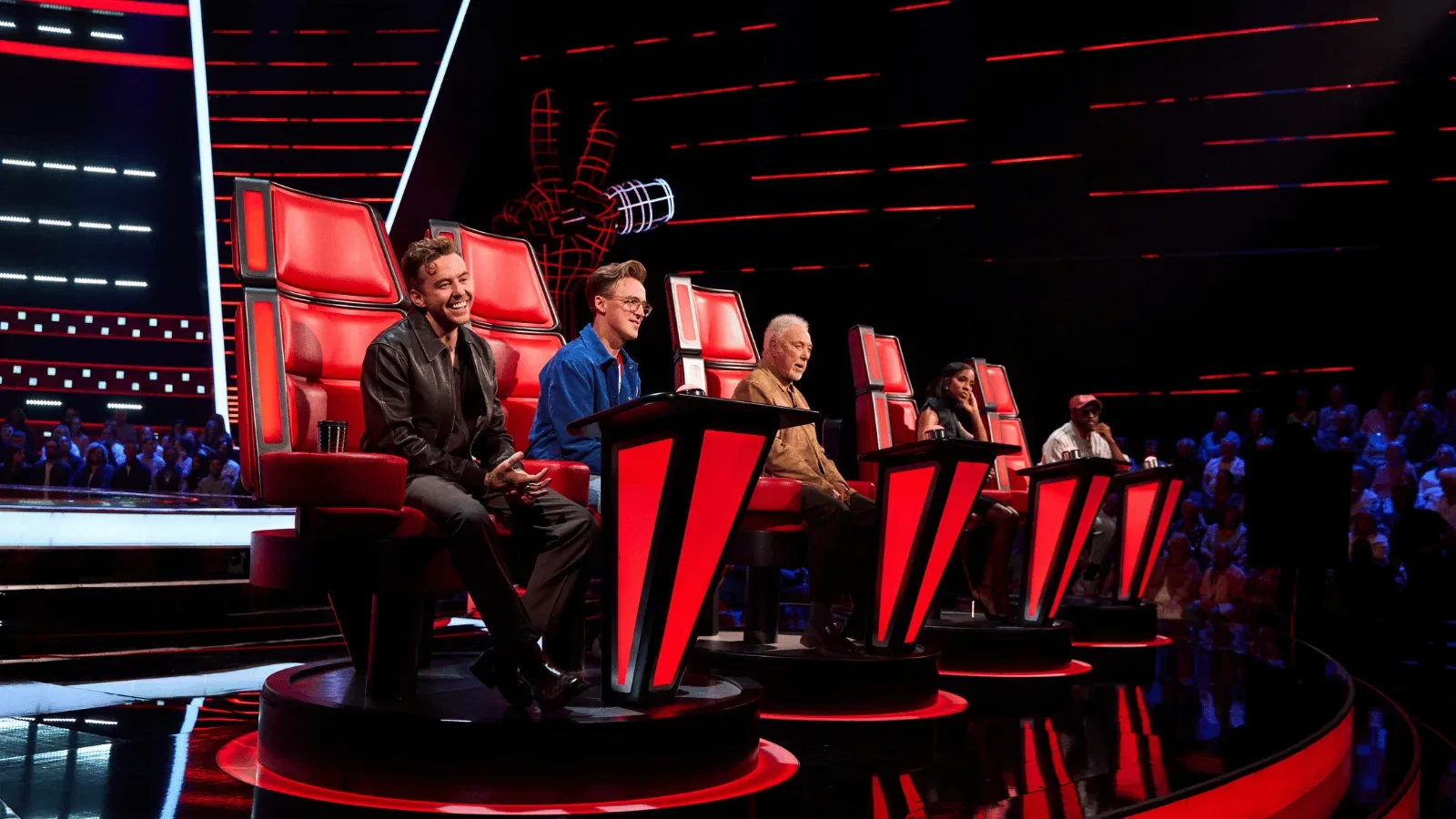 Five coaches sit in the iconic red chairs on a brightly lit singing competition stage. Red and black lighting surrounds the set, with a large hand-and-microphone logo visible in the background and a studio audience seated around the stage.