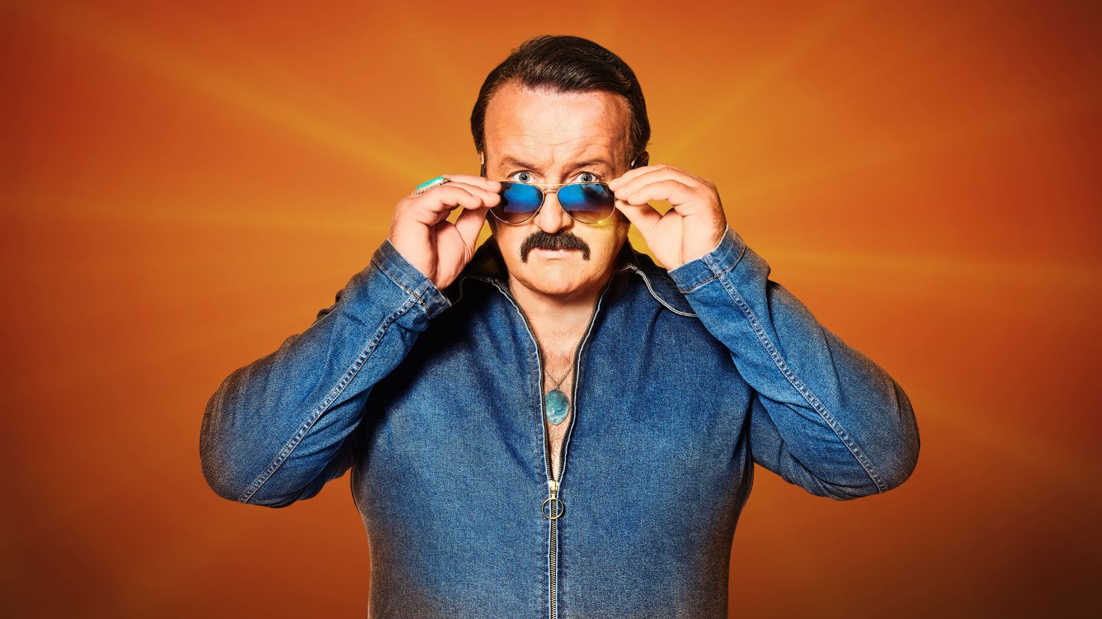 A man in the 80s wearing a denim outfit with slick hair and aviator sunglasses on stood against a plain orange background